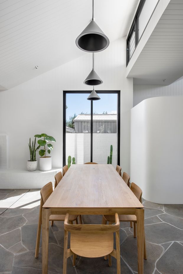 Bright contemporary dining room with a long light timber dining table and matching wooden chairs, set beneath three sculptural dark pendant lights. White vertical wall panelling and a high sloping ceiling create an airy volume, while large black-framed glazing overlooks a courtyard. The floor is finished in irregular dark grey stone slabs, with built-in white tiled seating or a low ledge and potted plants along the window.