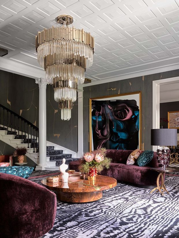 A dramatic, luxurious living room with a large tiered crystal-and-metal chandelier, dark charcoal walls, ornate white ceiling detailing, classical columns and staircase trim, deep burgundy velvet seating, a patterned monochrome rug, marble-toned coffee table, and bold abstract artwork. The room combines traditional architectural detailing with glamorous contemporary furnishings and rich jewel tones.