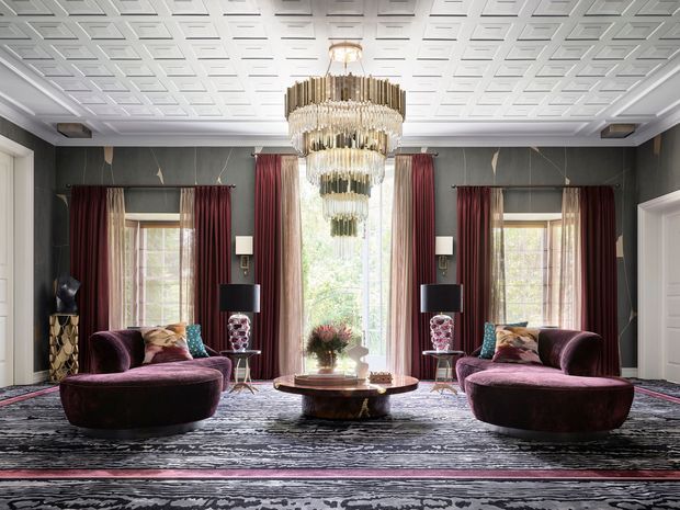 A highly styled, symmetrical formal living room with two deep burgundy velvet seating arrangements facing each other across a round coffee table. The room features dark charcoal wall panelling, tall windows with burgundy curtains and sheers, ornate crystal chandeliers, decorative lamps, patterned carpeting, and a striking geometric coffered ceiling. The overall composition is opulent, dramatic, and hotel-like.