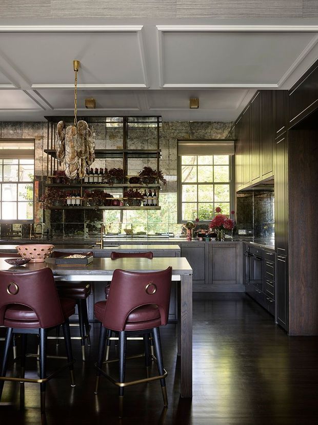 A sophisticated, richly detailed kitchen with a large central island and peninsula seating. The room features dark timber cabinetry, a dark polished timber floor, grey stone or concrete-look surfaces, mirrored and textured stone backsplash areas, and large black-framed windows. Burgundy leather bar stools with brass ring details sit at the island. The ceiling has decorative white coffered panelling, while an ornate clustered pendant light hangs above the island. Open shelving displays bottles and decorative objects.