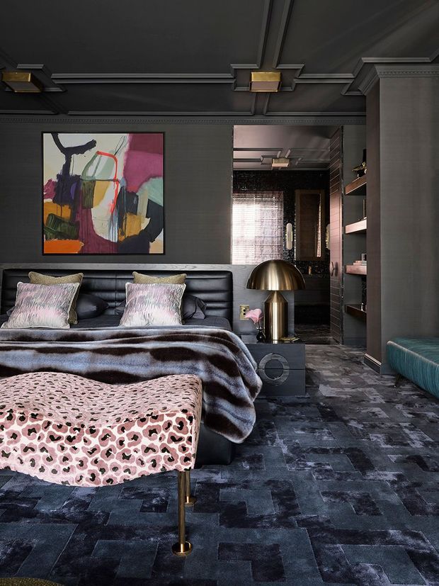 A richly styled, dark-toned bedroom with a black upholstered bed, layered patterned bedding, a pink animal-print bench at the foot, and a sculptural brass dome floor lamp. The room features charcoal wall panelling and coffered detailing, built-in open shelving, dark patchwork carpeting, a colourful abstract artwork, and a glimpse of a dark tiled adjoining dressing or bathroom area. The overall look is dramatic, luxurious, eclectic, and highly layered.