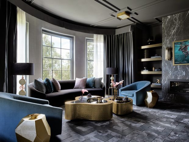 A dramatic, luxurious living room with a dark monochromatic palette, tall curved windows, layered black curtains, a black sofa with patterned cushions, blue upholstered armchairs, sculptural gold coffee tables, and a dark stone fireplace wall with built-in shelving. The floor is finished in dark grey patterned stone or tile, while a geometric ceiling light adds a contemporary focal point.