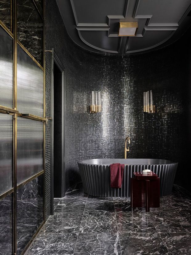 A dramatic, luxurious bathroom with a freestanding fluted black bathtub set against curved black mosaic-tiled walls. The room features dark marble flooring and lower wall panels, antique brass-framed translucent shower or partition glazing, brass wall sconces and tapware, and a decorative coffered dark ceiling with a central brass light fixture. A small dark wood side table and burgundy towel add contrast.