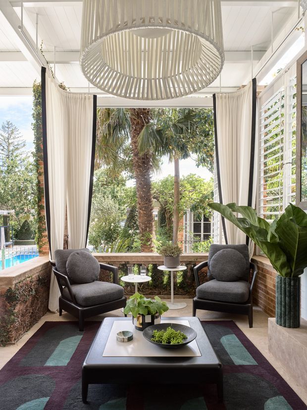 Bright, airy enclosed sunroom or lounge with large windows, plantation shutters, floor-to-ceiling curtains, exposed white ceiling boards, and views of a lush garden and pool. Two charcoal upholstered lounge chairs face a low dark coffee table over a geometric patterned rug, with potted plants and a large sculptural pendant light creating a relaxed indoor-outdoor atmosphere.