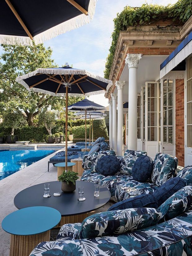 A luxurious outdoor poolside lounge beside a grand brick building. The terrace features patterned upholstered sofas and armchairs, round dark tables, blue accent tables, fringed navy umbrellas, formal white columns with ornate capitals, French doors, and lush planting surrounding a swimming pool. The setting has a refined resort-like atmosphere with strong blue, white, and green tones.