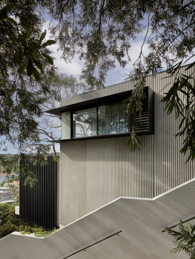 Exterior view of a modern multi-level residence on a sloped site, partially framed by overhanging trees. The building features dark vertical ribbed cladding, a light grey corrugated facade, large glazed windows with dark frames, and a steep concrete access ramp or driveway with a slim black handrail. The image emphasizes the building envelope and its relationship to the landscaped slope.
