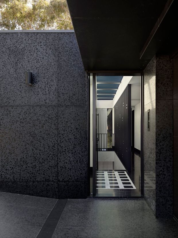 A dark, refined architectural entryway or covered external passage leading into a contemporary multi-level interior. Large dark speckled stone or concrete panels line the walls and floor, with black overhead soffits, slim black metal framing, a narrow linear drain, and a glazed opening revealing a stairwell or void beyond. Strong daylight enters through overhead glazing and creates geometric shadows on the floor. The composition emphasizes crisp rectilinear forms, deep charcoal tones, and a dramatic indoor-outdoor transition.