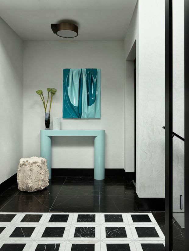 A refined residential entryway or hallway with white textured walls, black skirting and glossy black stone flooring. A pale aqua console table sits beneath a matching abstract artwork, accompanied by a sculptural light stone stool and a dark vase with dried stems. Geometric black-and-white patterned floor tiles define the foreground, while black-framed glazing or a doorway is visible on the right. The space has a bold, gallery-like contemporary aesthetic.