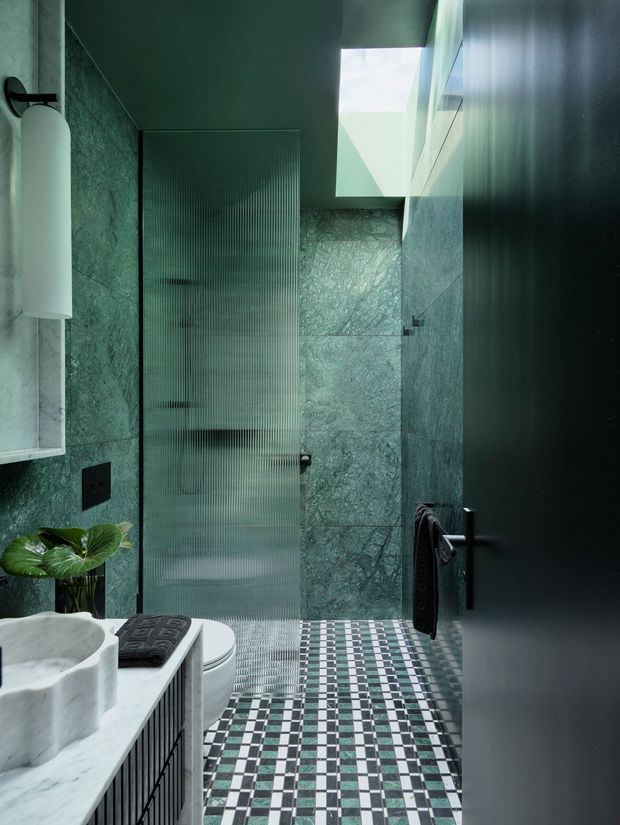 A compact, dramatically styled bathroom with green veined stone wall tiles, a patterned geometric mosaic floor, a sculptural white vessel basin on a dark ribbed vanity, a concealed toilet, ribbed translucent glass shower screen, and matte black fittings. A skylight brings in natural light, creating a moody yet refined atmosphere.