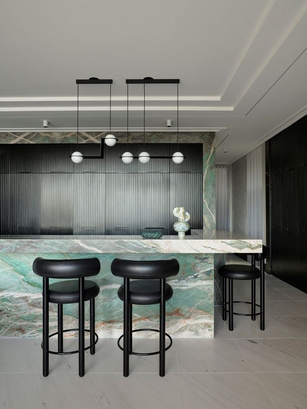 A sophisticated home bar with a long green-veined stone counter and matching stone-clad base, surrounded by black bar stools. The rear wall features dark vertical ribbed panelling, while sculptural globe pendant lights hang from a layered white ceiling. The palette is dramatic and refined, combining emerald and pale stone veining with black metal and dark joinery.