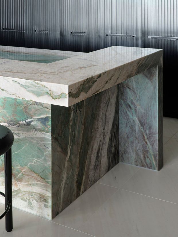 Close-up of a dramatic veined natural stone kitchen island or bar, with a thick waterfall-edge benchtop and matching stone-clad sides. The island is set against dark vertically ribbed wall panelling with slim linear wall lights, over a pale tiled floor; a small portion of a stool is visible at left.