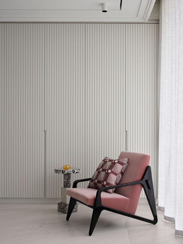 A refined interior corner featuring a pink upholstered mid-century-style lounge chair with black angular frame, positioned against a full-height light vertical fluted wall with concealed joinery doors. A small patterned side table and decorative object sit beside the chair. Sheer white curtains frame the right side, and the ceiling has layered recessed trim with a small black spotlight. The palette is soft and neutral with burgundy and yellow accents.
