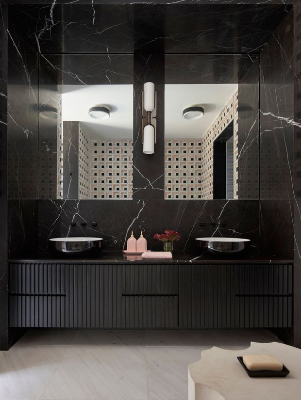A sophisticated contemporary bathroom vanity with a large double basin, fluted dark cabinetry, and dramatic black marble wall cladding with white veining. Two rectangular mirrors reflect a decorative breeze-block wall and ceiling lights. The room includes wall-mounted fixtures, sculptural pendant lighting, pale stone flooring, and a light stone foreground counter or bench.