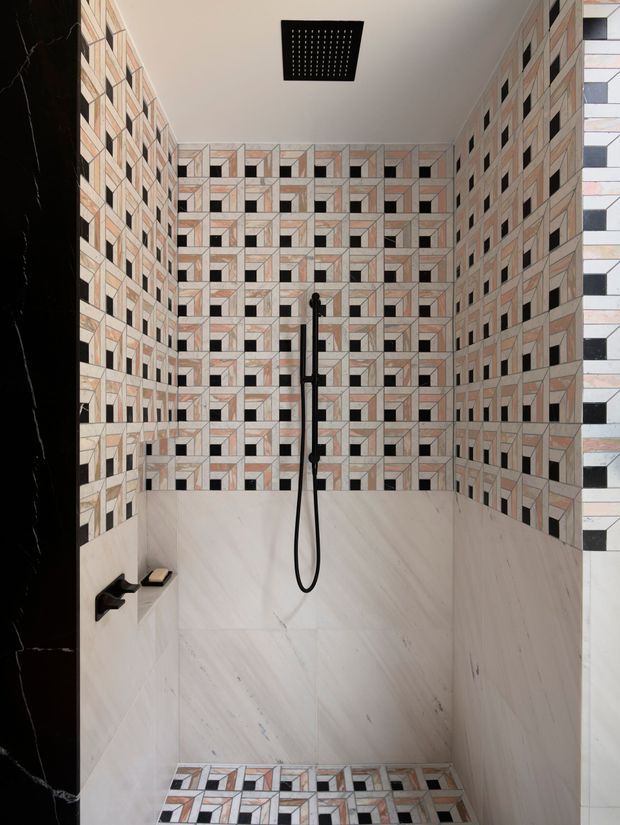 A contemporary walk-in shower enclosure with a striking geometric patterned tile feature wall and matching tiled floor border. Large pale marble-look wall tiles cover the lower walls, while matte black shower hardware, controls, and a square ceiling rain shower provide contrast. Dark marble-look wall trim frames the opening.
