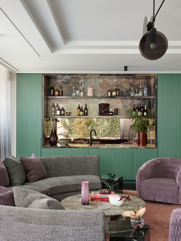 A stylish living room with a large curved grey sectional sofa and mauve textured armchairs arranged around a round stone coffee table. The room features a dramatic built-in bar with mirrored or reflective stone shelving, bottles, glassware, and a sink, framed by green vertical fluted wall panelling. A sculptural dark pendant hangs from a recessed coffered ceiling, while large glazing or reflective surfaces bring in greenery and daylight. The palette is moody and refined, combining green, grey, mauve, dark stone, and metallic accents.