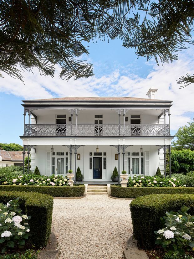 Front-on view of a grand two-storey heritage-style residence with a symmetrical white facade, full-width decorative cast-iron verandah and balcony, dark entrance door, shuttered windows, hipped roof, and a formal landscaped garden with clipped hedges, gravel paths, topiary, and hydrangeas.