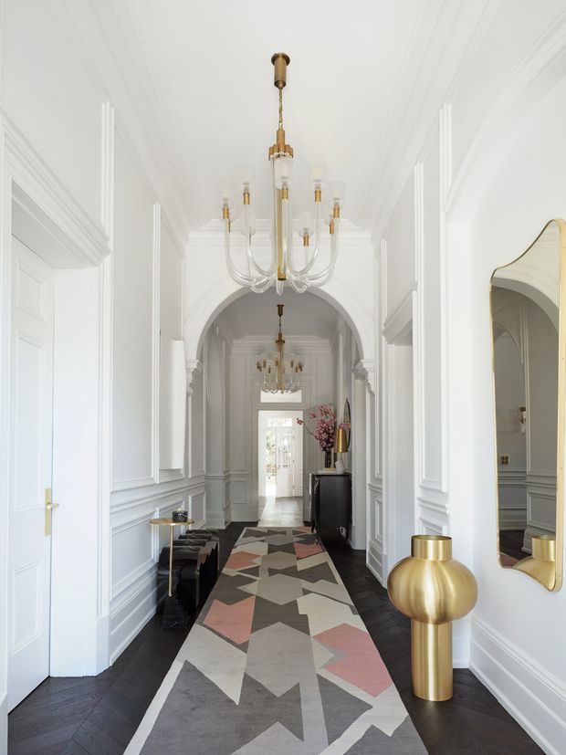 A bright, elegant residential hallway with white architectural mouldings, paneled walls, decorative arched openings, dark timber flooring, and multiple brass-and-glass chandeliers. A geometric patterned runner extends through the corridor, with a console, sculptural brass lamp, mirror, and dark cabinetry adding refined decorative accents.