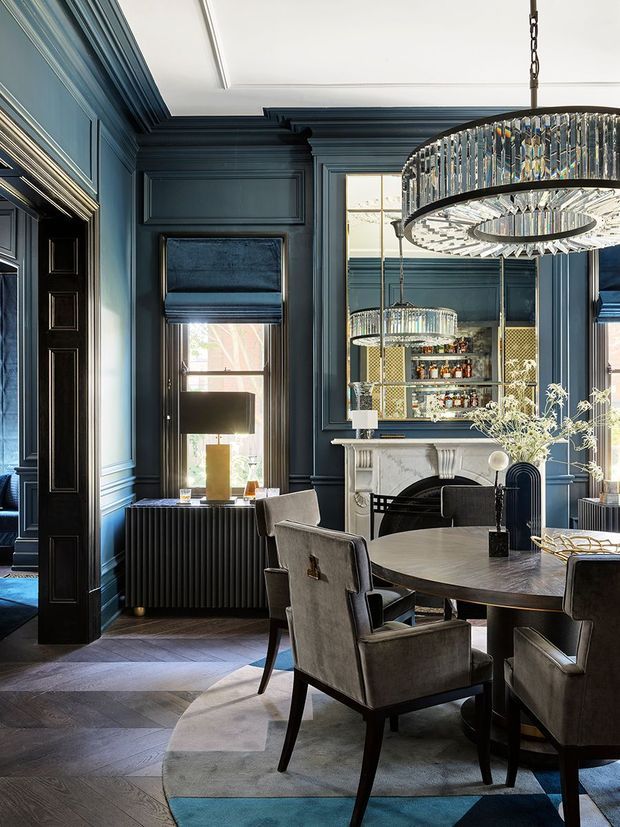 A sophisticated dining room with deep blue panelled walls, ornate trim, a dark herringbone timber floor, and a large round dining table surrounded by upholstered chairs. A marble fireplace sits beneath a mirrored bar or display cabinet, while statement crystal chandeliers add a luxurious focal point. The room has tall windows with dark Roman blinds and a refined, dramatic atmosphere.