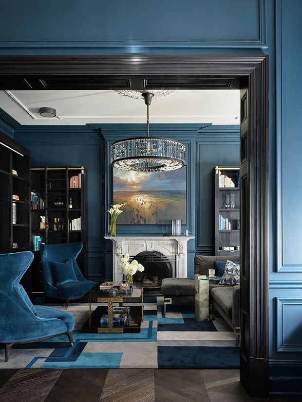A richly detailed, dark blue living room viewed through a wide decorative doorway. The room features deep navy-blue wall panelling and matching built-in bookcases, a white carved fireplace with a landscape artwork above it, a large crystal chandelier, velvet lounge chairs, a sectional sofa, a glass coffee table, and a geometric blue, grey, and cream rug over dark herringbone timber flooring. The space has a dramatic, refined, traditionally styled interior with strong layered moulding and high-contrast finishes.