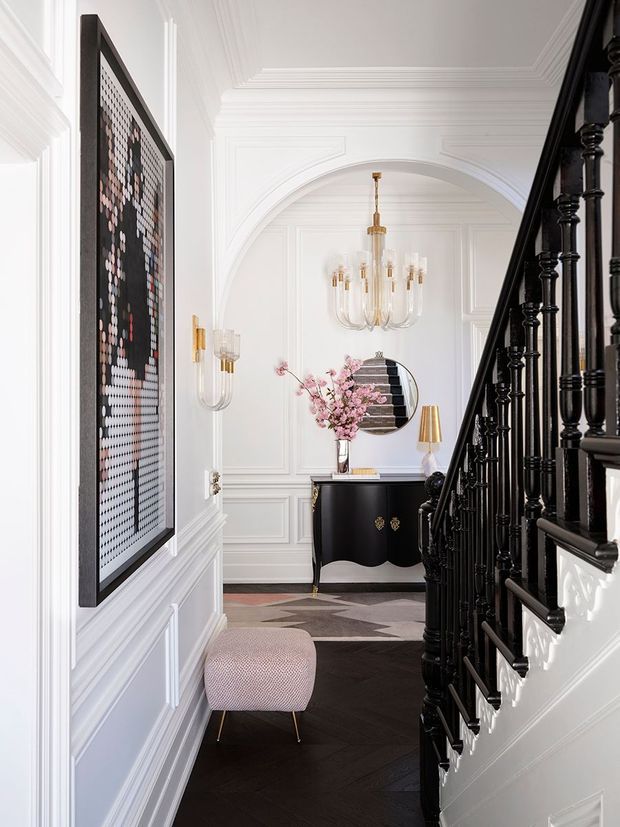 Elegant, bright residential hallway and stairway with elaborate white decorative wall panelling, moulded ceiling trim, and a large arched opening. A glossy black staircase balustrade dominates the right side, while a black console with gold details, round mirror, sculptural chandelier, wall sconces, and pink floral arrangement create a refined, art deco-inspired entry composition. Dark geometric flooring contrasts with the white architectural shell.