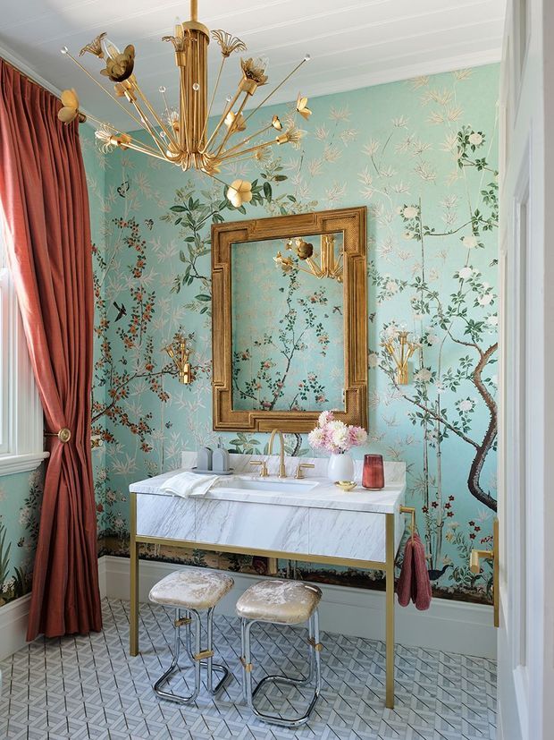 A highly decorative bathroom vanity area featuring a white marble-topped console with brass legs, a large ornate framed mirror, patterned botanical wallpaper, brass floral chandelier and sconces, deep burgundy drapery, upholstered stools, and geometric patterned floor tiles. The room has a richly layered, glamorous, vintage-inspired aesthetic.