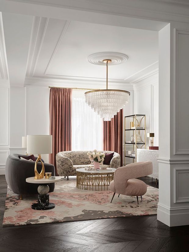 Elegant formal living room with white decorative wall panelling, ornate ceiling mouldings and a central chandelier. The room features a dark herringbone timber floor, large muted floral-patterned rug, curved upholstered sofas and armchairs, a round glass-topped coffee table with a metal base, sheer curtains with dusty rose drapes, and a slim open shelving unit. The overall look is refined, luxurious, and contemporary with classic architectural detailing.
