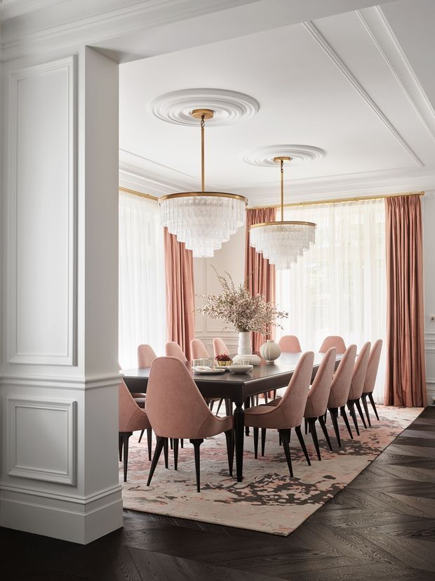Elegant formal dining room viewed through a wide opening, featuring a large dark rectangular dining table surrounded by blush-pink upholstered chairs, two tiered crystal pendant chandeliers, ornate concentric ceiling medallions, wall panel mouldings, floor-to-ceiling windows with sheer and dusty-rose curtains, and a patterned area rug over dark herringbone timber flooring. The room has a refined, warm, high-contrast aesthetic with classical detailing and contemporary furniture.