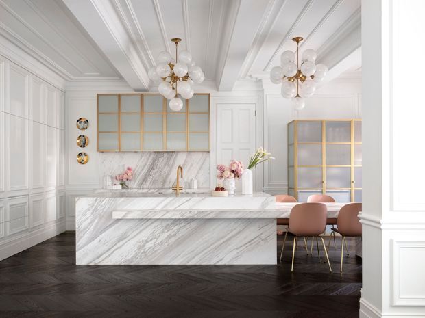 Elegant open-plan kitchen and dining area with a large veined marble island, white paneled walls and coffered ceiling, dark herringbone timber flooring, brass-framed glazed cabinetry, and sculptural globe pendant lights. Blush upholstered dining chairs and floral arrangements add soft colour to the refined, high-contrast scheme.