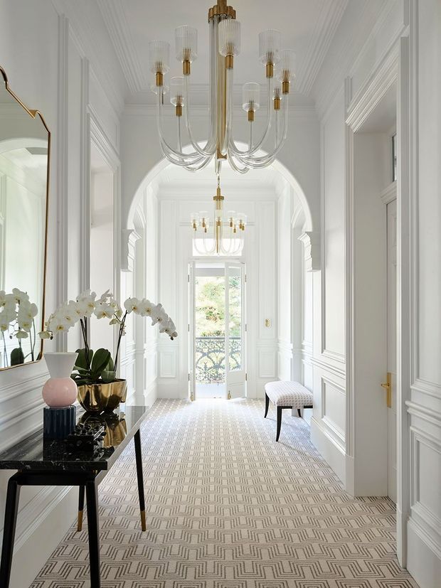 A bright, elegant residential hallway or entry gallery with extensive white wall panelling, decorative mouldings, arched openings, and a patterned geometric carpet. Multiple sculptural chandeliers with clear glass arms and brass/gold accents hang from the high ceiling. A black console table with orchids, decorative objects, and a large mirror occupies the left side, while a small upholstered bench sits along the right wall. Glazed French doors at the far end open toward greenery and a decorative balcony or exterior area.