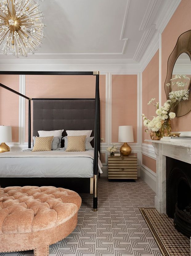 Elegant, richly detailed bedroom featuring a black four-poster canopy bed with a tufted upholstered headboard, pale bedding, and patterned accent pillows. The room has blush-pink wall panels with elaborate white mouldings, a marble fireplace with a dark inset, a sculptural wavy mirror, brass-toned bedside lamps and chest, a crystal starburst chandelier, and a large blush velvet tufted ottoman. A geometric patterned carpet covers the floor, creating a refined, glamorous and highly layered interior.