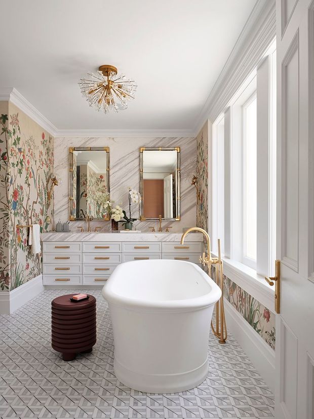 A bright, elegant bathroom with a freestanding white bathtub positioned beside tall windows, a double vanity with marble-look surfaces, twin framed mirrors, patterned botanical wallpaper, geometric patterned floor tiles, and polished brass fixtures. A decorative gold chandelier adds a refined focal point.