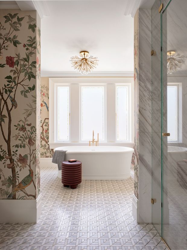 Elegant, bright bathroom featuring a freestanding white soaking tub beneath tall narrow windows, patterned geometric floor tiles, botanical floral wall covering, and a glass-enclosed marble shower on the right. A sculptural gold starburst pendant, brass fixtures, and a small burgundy ribbed stool add refined decorative accents.