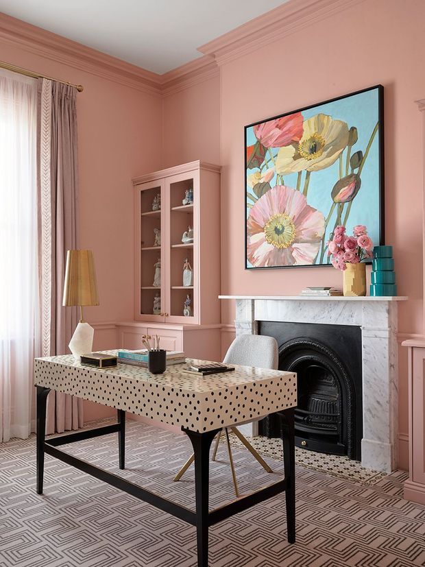 A refined, colourful home office with dusty pink walls, ornate ceiling cornices and wall panelling, tall curtained windows, a patterned geometric carpet, and a black-framed marble fireplace. A distinctive spotted desk with black legs sits in the foreground, accompanied by a light upholstered chair. Built-in pink cabinetry displays decorative objects, while a large floral artwork and sculptural table lamp add bold character.