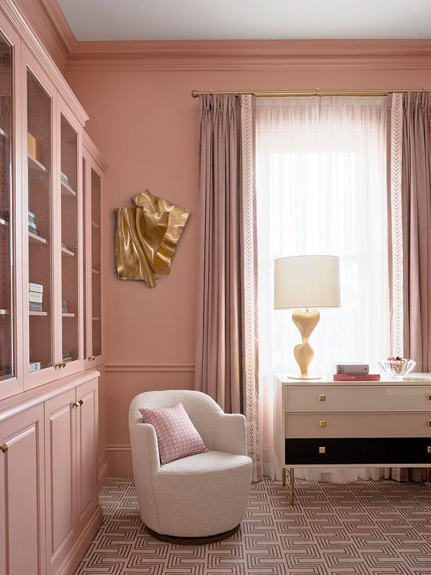 A refined, compact sitting or study area in a soft blush-pink palette. The room features extensive built-in glass-front cabinetry with classic panelled joinery, decorative wall moulding, a patterned geometric carpet, a sculptural upholstered armchair, and a vintage-style chest beneath a tall window. Sheer curtains and taupe-pink drapery soften the daylight, while brass and gold accents add warmth and polish.
