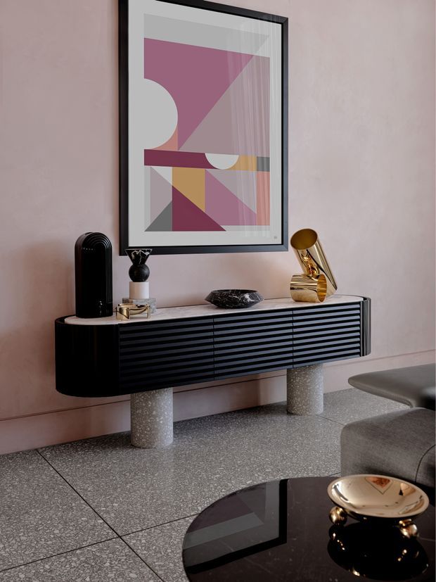 A refined contemporary living room vignette featuring a wall-mounted black fluted sideboard with cylindrical terrazzo supports, a pink-toned abstract geometric artwork in a black frame, sculptural decor, and a polished gold table lamp. The room has a pink plaster wall, terrazzo flooring, and a partially visible grey sofa with a glossy black coffee table in the foreground.