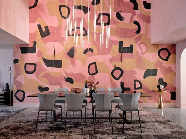 A refined contemporary dining area with a long dining table and upholstered grey dining chairs, set against a large expressive abstract mural in pink, peach, mauve and black. Sculptural pendant lighting hangs above the table, while minimalist shelving, decorative objects and polished finishes create a sophisticated, gallery-like interior.