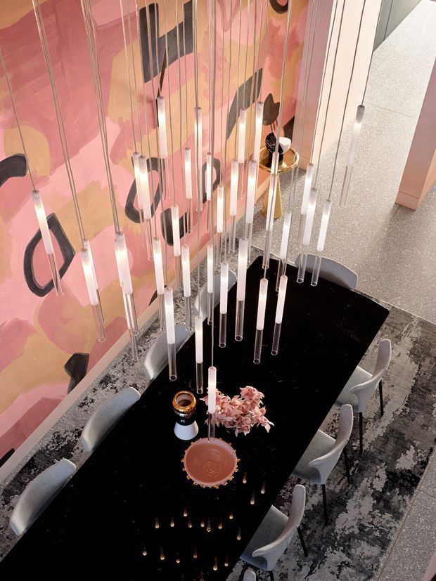 Overhead view of a refined dining area with a long glossy black dining table surrounded by upholstered grey chairs. A dramatic cluster of suspended linear pendant lights hangs above the table, while the surrounding walls feature a bold abstract pink, peach, black and cream mural. A patterned grey rug and sculptural table decor complete the space.