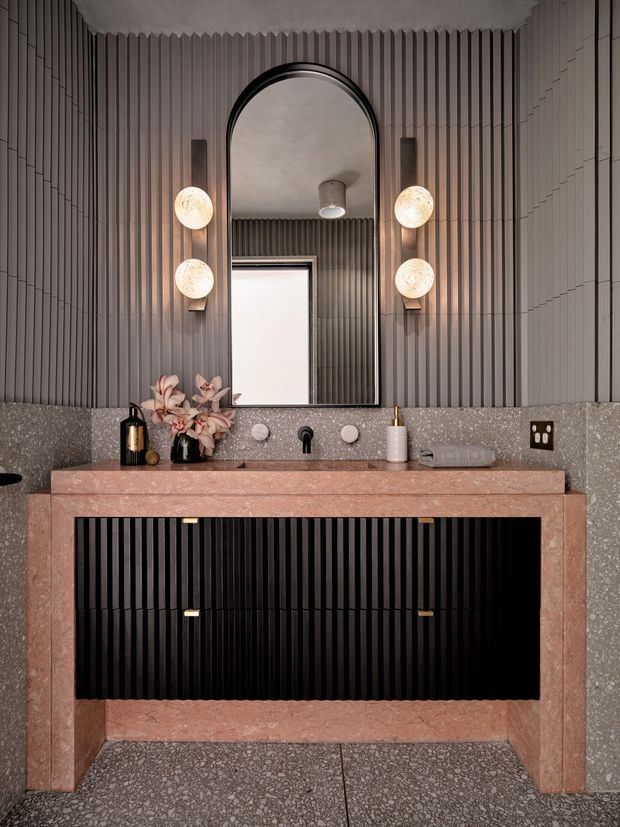 A refined contemporary bathroom vanity viewed head-on. The space features a wide double vanity with a pink-toned stone benchtop and thick framed surround, black fluted cabinetry below, and a matching speckled stone backsplash and floor. Tall vertically ribbed wall panelling creates a textured backdrop, with an oversized arched mirror, paired globe wall sconces, twin basins and dark tapware. The palette is warm, muted and luxurious, with black, dusty pink stone and grey terrazzo-like surfaces.