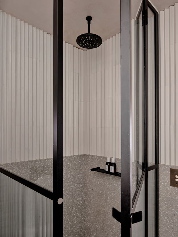 A contemporary shower enclosure viewed through a black-framed glass door. The bathroom features vertically fluted light wall panels, a terrazzo-style lower wall and floor finish, a large round black overhead shower, and a small built-in shelf with toiletries. A mirror or reflective glass panel is visible on the right.