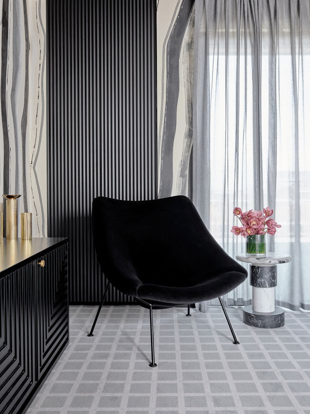 A refined interior seating vignette featuring a sculptural black lounge chair with slender metal legs, positioned beside a black fluted console cabinet. The room has a dramatic vertically ribbed black feature wall, pale patterned carpet, sheer floor-to-ceiling curtains, and a small round side table with a marble-like top and cylindrical pedestal. Metallic decorative vessels and a vase of pink flowers add accents.