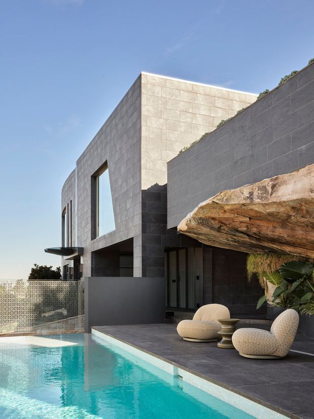 Contemporary multi-level residence built into a sloping site, featuring large-format dark and pale stone-clad walls, tall glazing, a projecting rock-like roof or canopy, and a turquoise swimming pool with a tiled privacy screen. Outdoor seating occupies the pool terrace, with integrated planting and distant urban views.