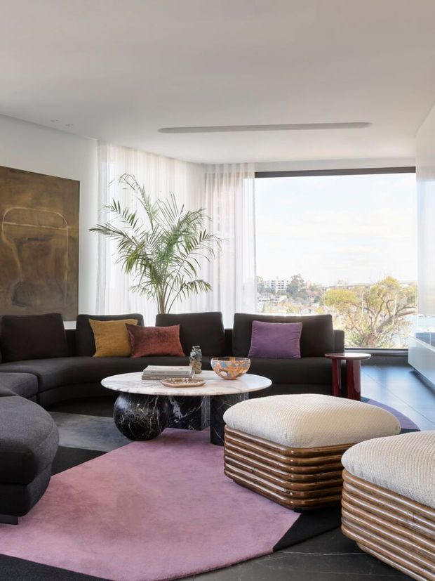 Bright, sophisticated living room with floor-to-ceiling sheer curtains and a large picture window overlooking an urban landscape. The room features a deep charcoal curved sectional sofa with colourful cushions, a round white marble coffee table with a dark sculptural base, two ribbed upholstered ottomans, a sculptural indoor palm, contemporary artwork, and a large mauve-and-black area rug. The overall look is polished and modern with strong curved forms and an airy outlook.