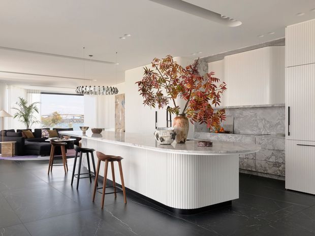 A bright, refined open-plan kitchen and living area with a large curved island, white vertically fluted cabinetry, a pale veined marble backsplash and benchtop, and dark stone-look flooring. Timber bar stools and dark lounge furniture contrast with the light joinery. A dining and living zone is visible beyond the island, with abundant glazing and sculptural pendant lighting.