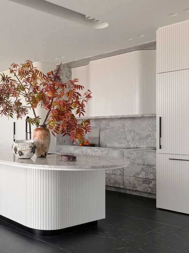 A refined contemporary kitchen featuring a curved island with vertically fluted white cabinetry, a pale stone benchtop, tall fluted storage cabinetry, and dramatic grey-veined stone wall cladding and splashback. The space has dark stone flooring, recessed ceiling lighting, sculptural integrated ceiling detailing, and warm botanical styling on the island.