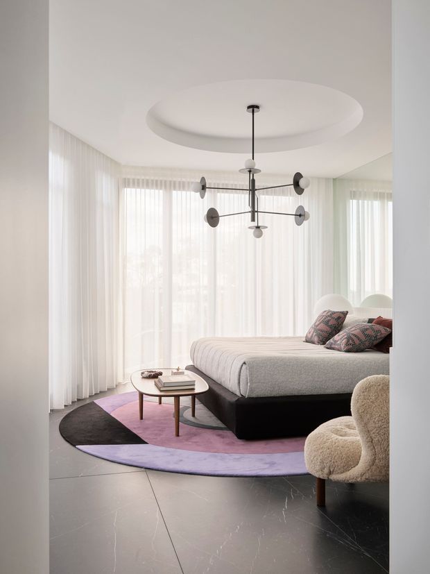A bright, contemporary bedroom with floor-to-ceiling sheer curtains, a large upholstered bed, sculptural black-and-white pendant light, curved ceiling feature, round coffee table, boucle armchair, and a layered geometric rug in black, lavender, and pink. The room has a polished dark stone-look floor and a soft neutral palette with colourful accents.