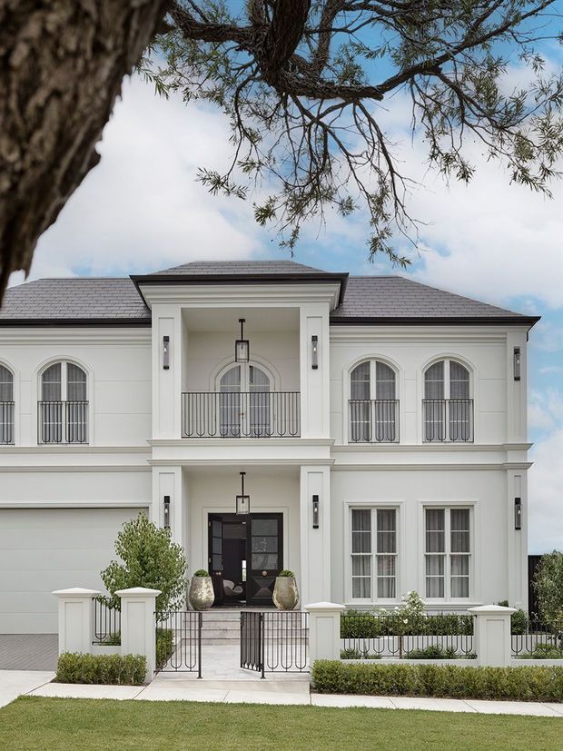 Front exterior view of a symmetrical two-storey white rendered residence with a hipped grey tiled roof, classical proportions, arched windows, decorative mouldings, Juliet balconies, black metal balustrades, and a formal entry gate. A landscaped front lawn and mature tree frame the facade.