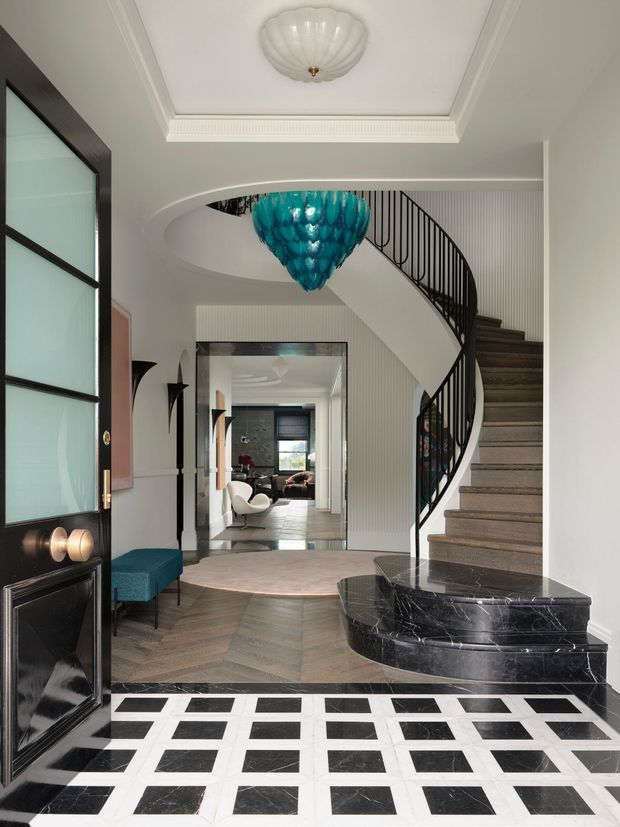Elegant residential entry hall with a dramatic curved staircase, black metal balustrade, sculptural turquoise pendant light, decorative ceiling mouldings, black-and-white geometric marble flooring, and a rounded black marble stair base. A teal upholstered bench sits beside the entry, with a living room visible through the opening beyond.