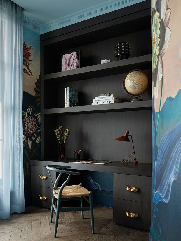 A compact built-in home office nook with dark timber shelving and cabinetry, a desk, sculptural chair, and warm red desk lamp. The deep blue-black joinery is framed by vivid botanical wallpaper, with decorative objects including books, a globe, flowers, and artwork. Gold drawer pulls and a herringbone timber floor add refined detail, while sheer curtains soften the space.
