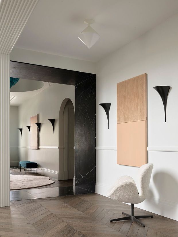 A refined interior vignette featuring a large black marble-framed mirror, pale moulded walls with dado detailing, sculptural black wall sconces, a geometric pendant light, and two-tone timber wall art. A boucle swivel chair sits on a warm herringbone timber floor, while the mirror reflects an arched doorway, curved furnishings, and an adjacent seating area.