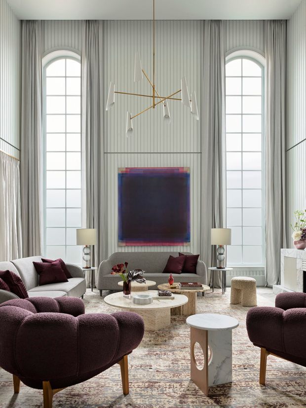 A refined, double-height living room with tall arched grid windows, full-height pleated curtains, vertically panelled walls, and a sculptural contemporary chandelier. The room is furnished with curved light-grey sofas, deep burgundy boucle armchairs and cushions, round stone coffee tables, side tables, lamps, and a large muted patterned rug. A large abstract artwork forms the focal point above the central sofa. The palette is soft grey, white, burgundy, and warm brass, creating a luxurious and dramatic yet inviting interior.